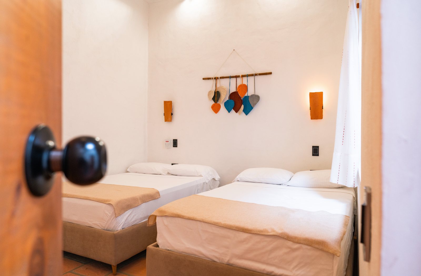 Habitación con dos camas semidobles, apliques de madera y un móvil artesanal sobre el cabecero